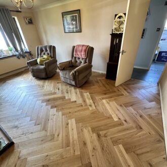 Warm living room with natural oak herringbone flooring, two recliners, a wall clock and an open doorway leading to a hallway.